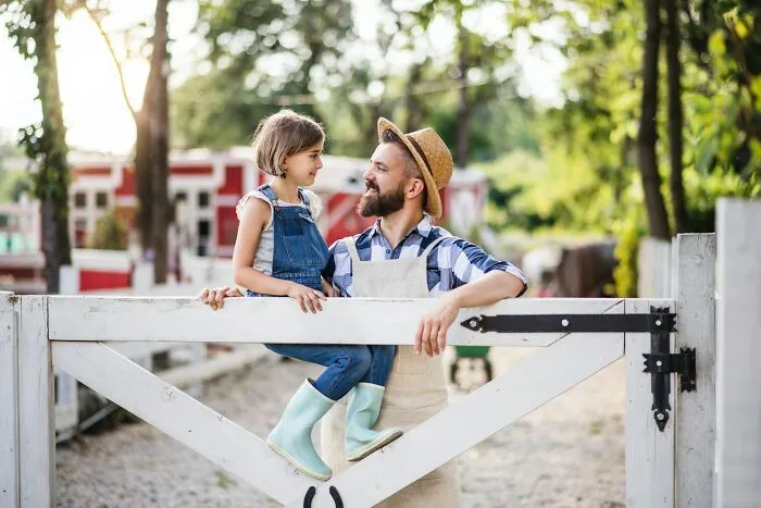 A dad wearing a straw hat and plaid shirt with his daughter on a fence, discussing advice for raising daughters.