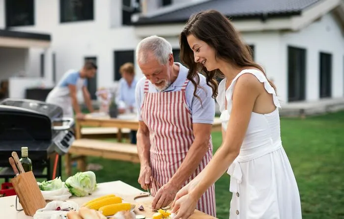 A dad in an apron and his daughter smiling while preparing vegetables for a barbecue. Women giving dads advice on raising daughters.