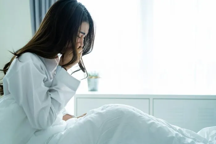 A distressed woman in a white shirt sits on a bed, holding her head, reflecting patient neglect in emergencies.