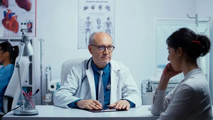 A doctor in a white coat and glasses sitting at a desk across from a female patient, who looks concerned. Patients being ignored.