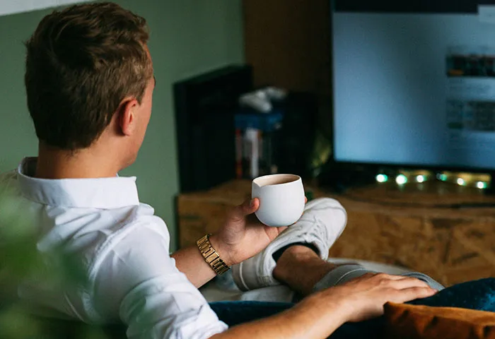 Man relaxing with a drink at home, watching TV and reflecting on harmless but effective ways to get revenge on jerks.