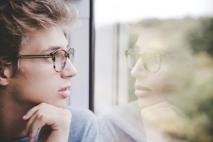 A young man with glasses looks thoughtfully out a window, reflecting on family secrets.