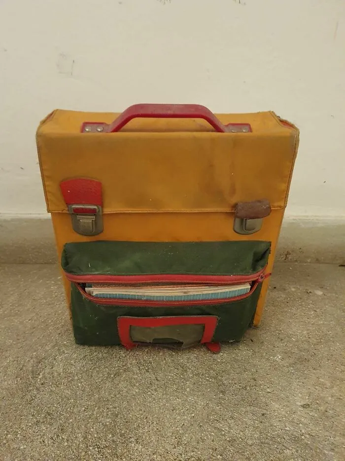 Old yellow and green bag with red handle and latches, showing pareidolia as it resembles a face, on a concrete floor.