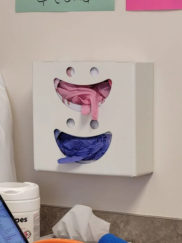 Smile-shaped glove dispenser on a wall showing pareidolia with pink and blue gloves inside.
