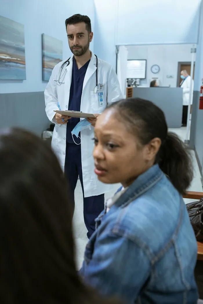 A doctor with a clipboard and a surgical mask looks on as a patient is ignored in emergencies, looking concerned.