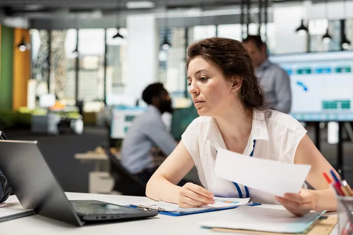Woman at a desk with a laptop, reviewing documents. Absurd Rules and workplace policies are common topics.