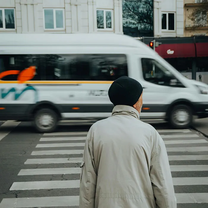 Man wearing a hat standing at a crosswalk while a white van passes by, illustrating safety tips for pedestrians.