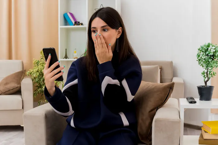 Young woman sitting on a sofa looking shocked at her phone, capturing a moment of infidelity getting exposed.