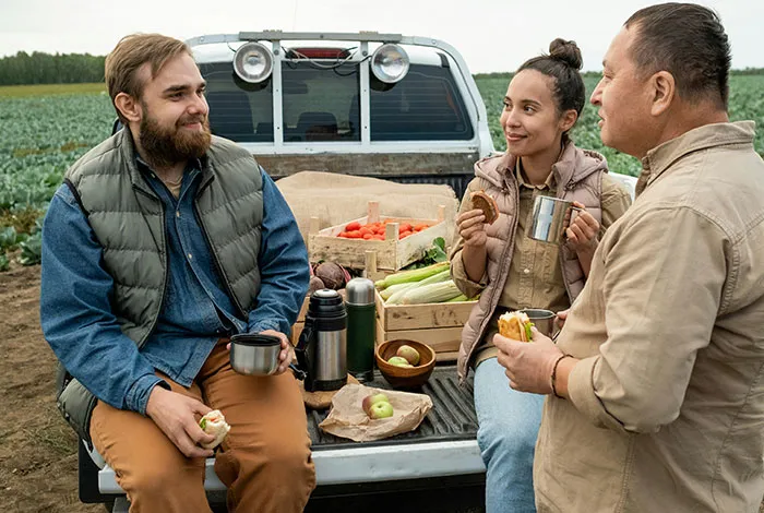 Three people showcase intelligence during a farm break, enjoying lunch on a truck tailgate with fresh produce.