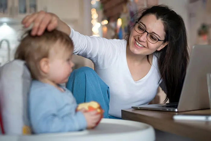 A smart, smiling woman in glasses works on a laptop, gently touching a baby's head, highlighting a moment of natural intelligence.