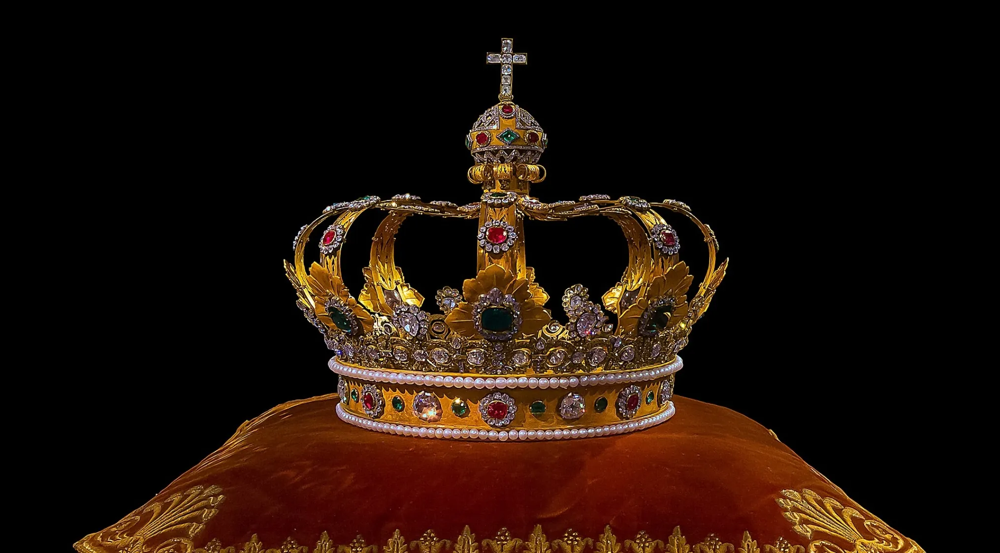 An ornate gold crown adorned with many gems, including rubies, emeralds, and pearls, resting on a velvet pillow. Australian Citizenship.