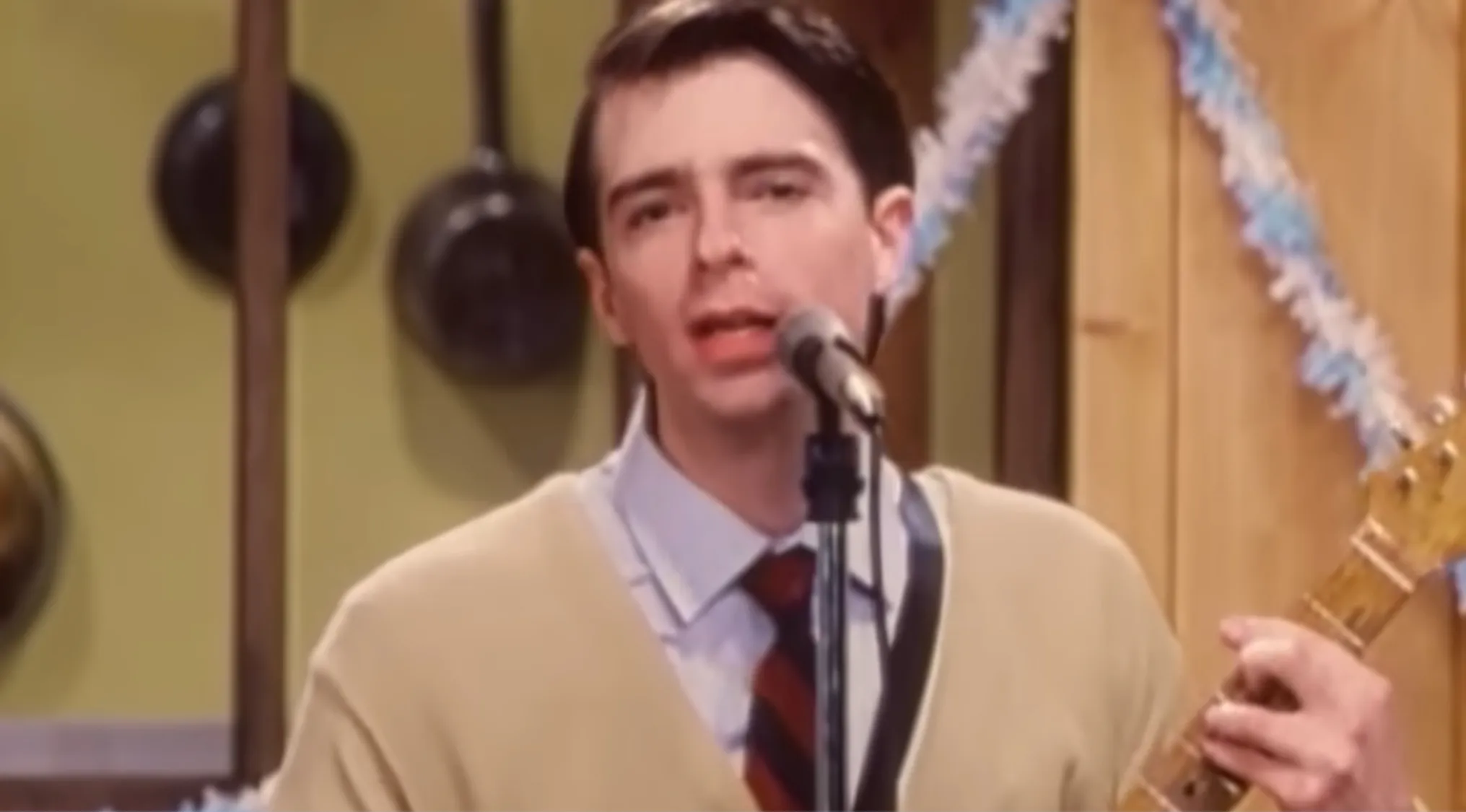 A male lead singer in a tan cardigan and striped tie, playing a guitar and singing into a mic. Music challenge: Name 30 bands.