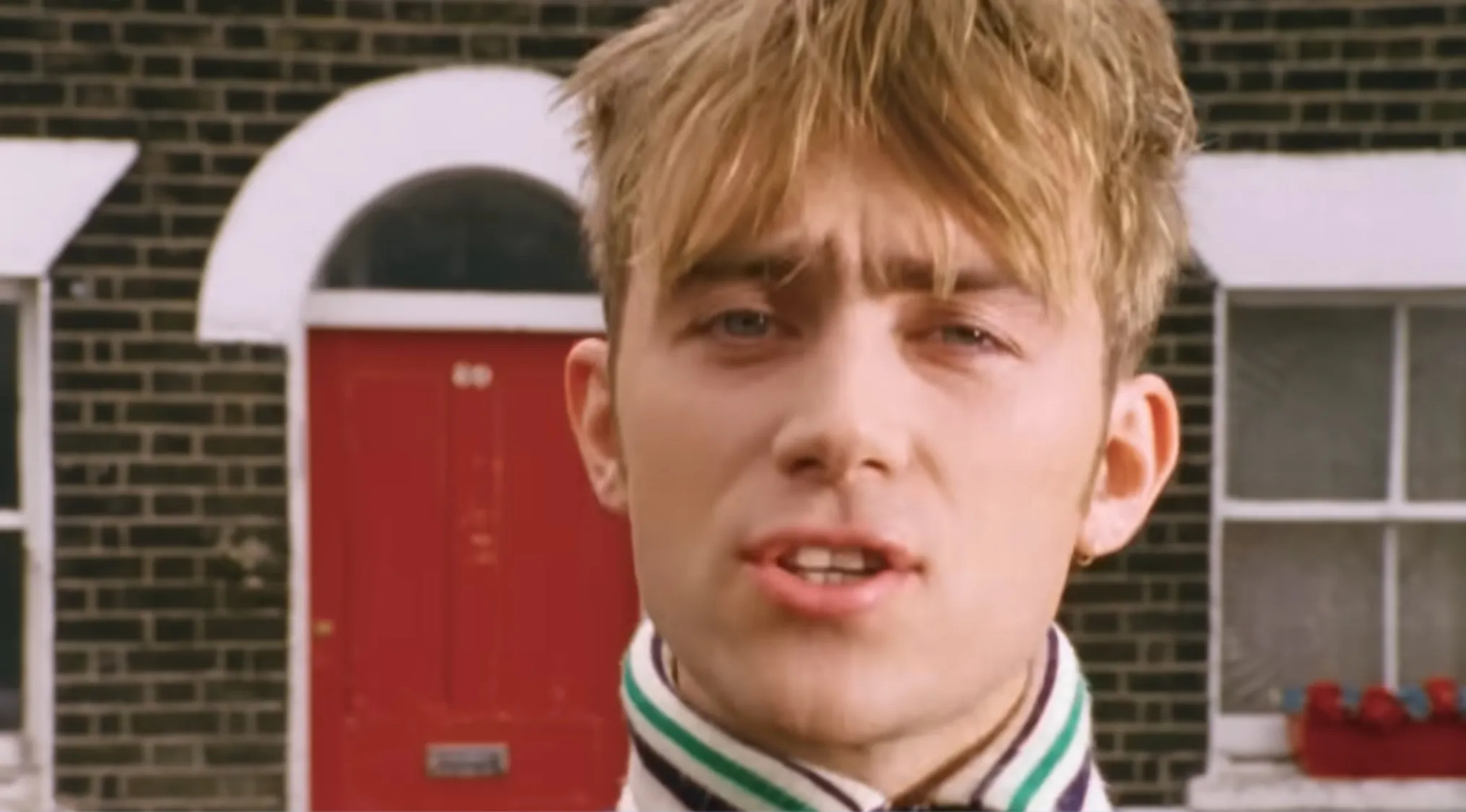 Damon Albarn, lead singer of a band, with blond hair, blue eyes, and striped collar, in front of a red door.