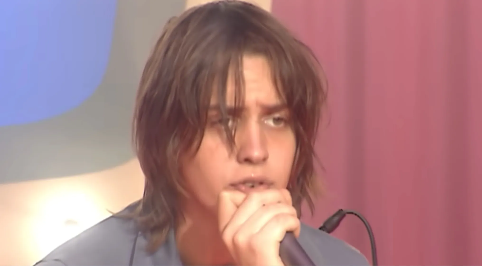 Julian Casablancas, frontman, singing into a microphone with his hand covering his chin, messy brown hair, in a music challenge setting.
