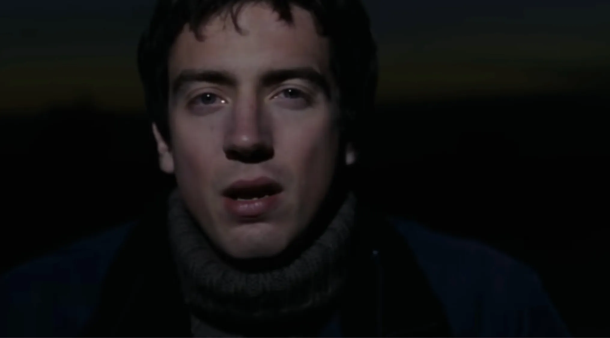 A close-up of a young male vocalist with dark hair, wearing a dark blue jacket and a grey roll-neck. Music challenge.