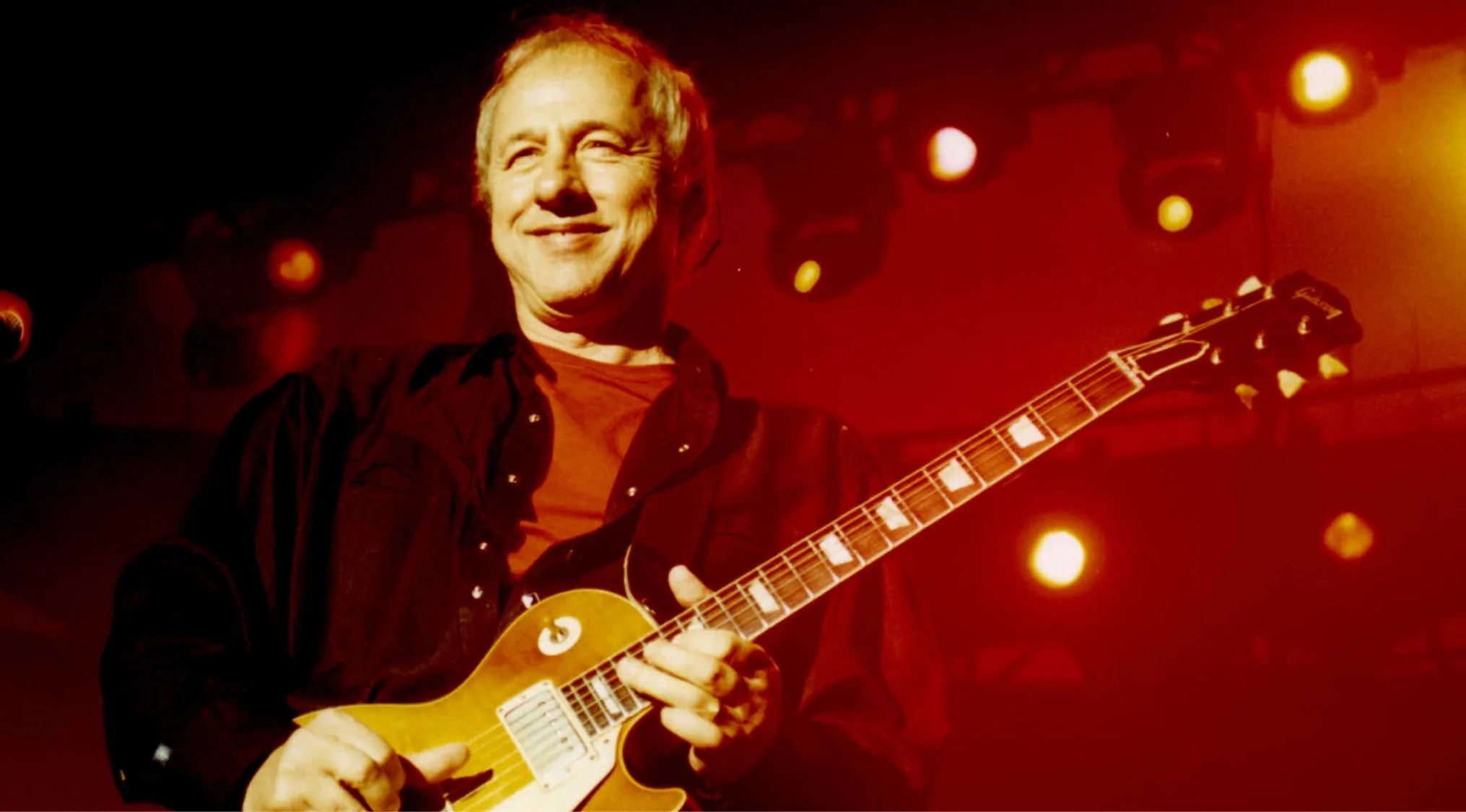 Mark Knopfler, frontman, playing guitar live on stage under red lights. Music challenge to name 30 bands.