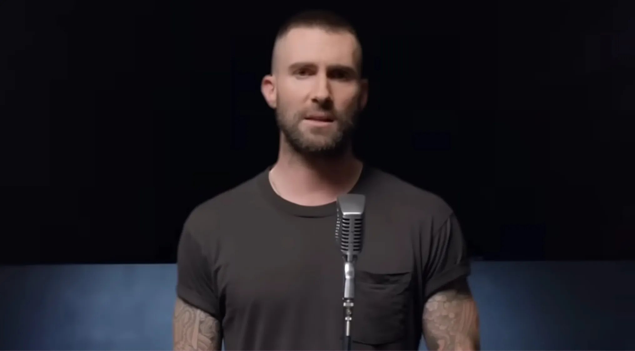 Adam Levine, lead singer with a beard and buzz cut, wearing a t-shirt, standing at a microphone for a music challenge.