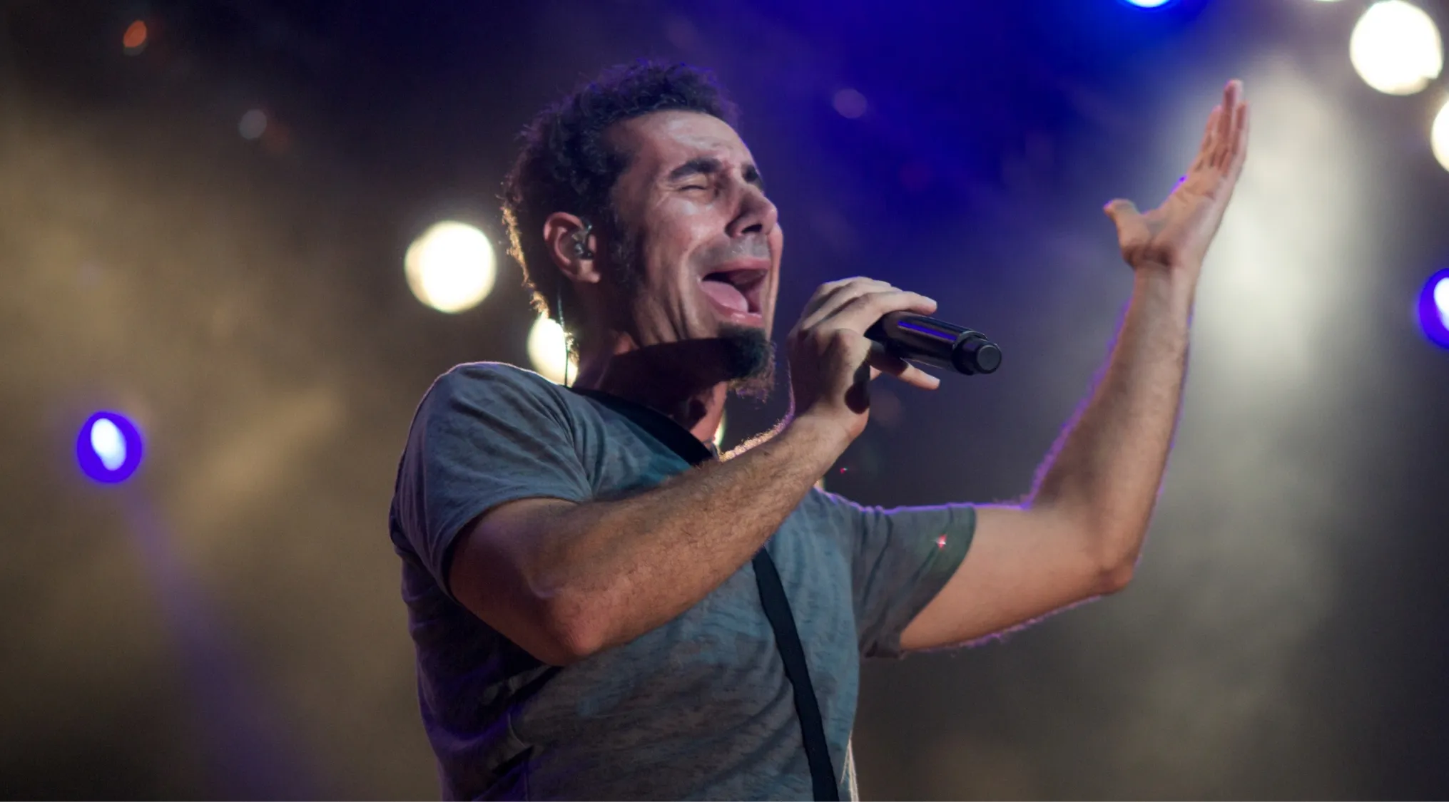 Serj Tankian, lead vocalist, performing intensely on stage with a microphone, lit by blue and white lights. Music challenge.