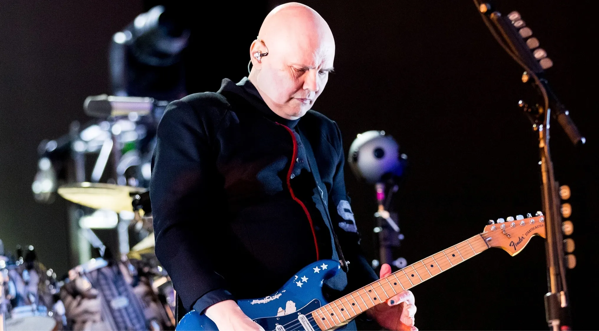 A bald lead singer on stage, focused on playing a blue electric guitar. Identify bands from lead singers.