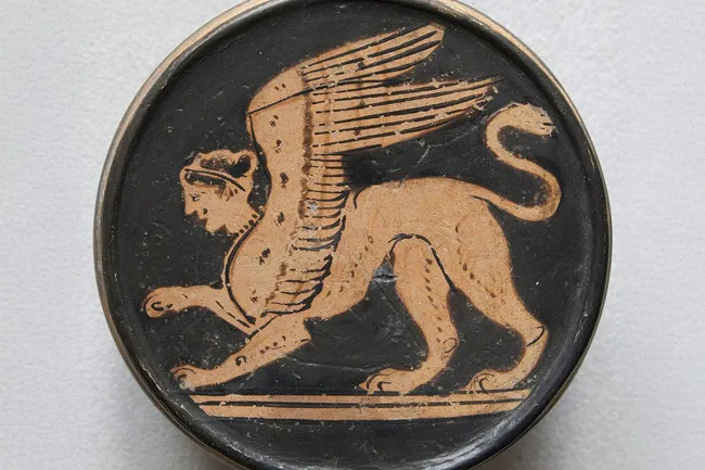An ancient Greek Sphinx depicted on a round, black pottery shard, in a mythical creature design.
