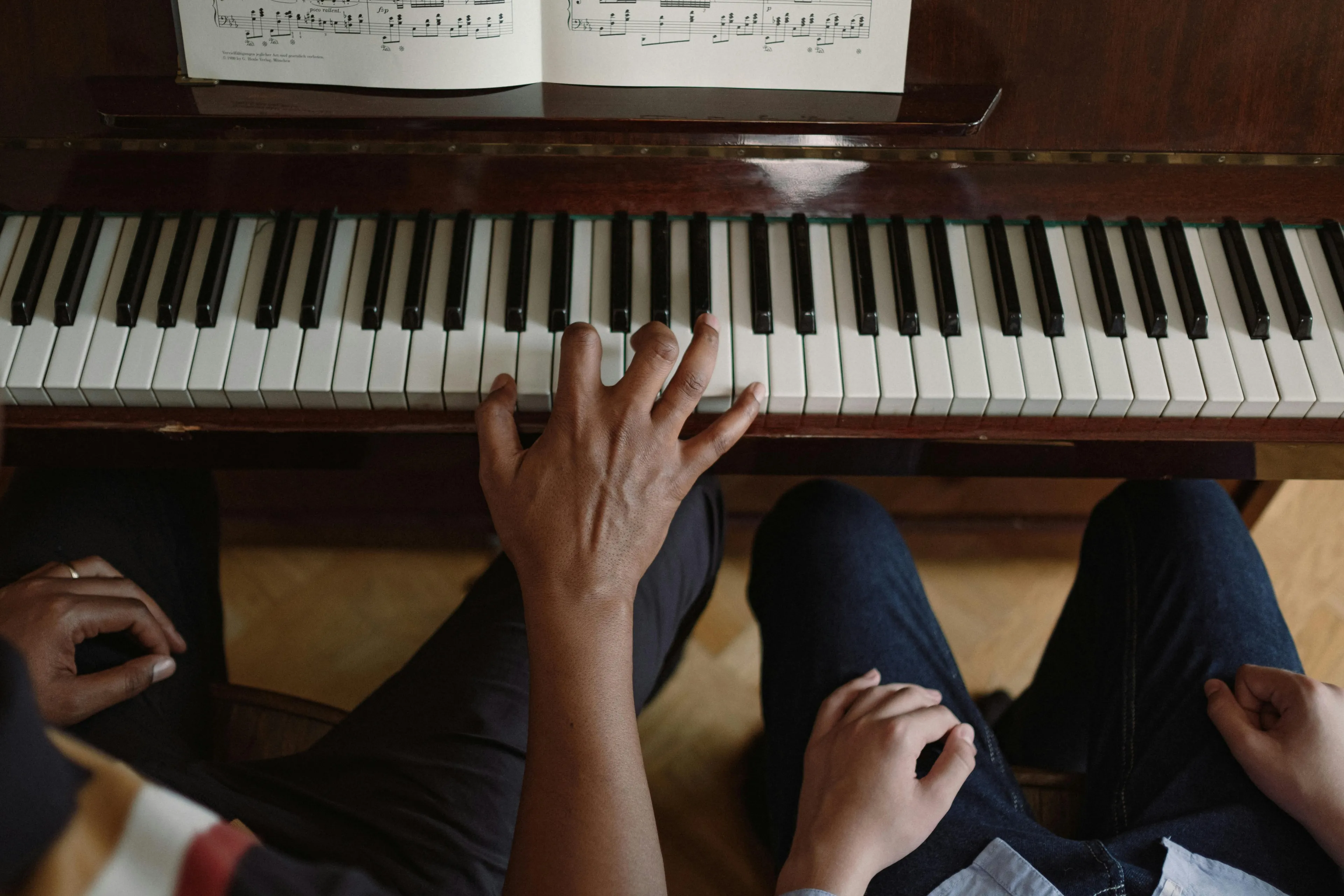 Overhead shot of two people sitting at a piano with sheet music; one hand plays keys, highlighting music education.