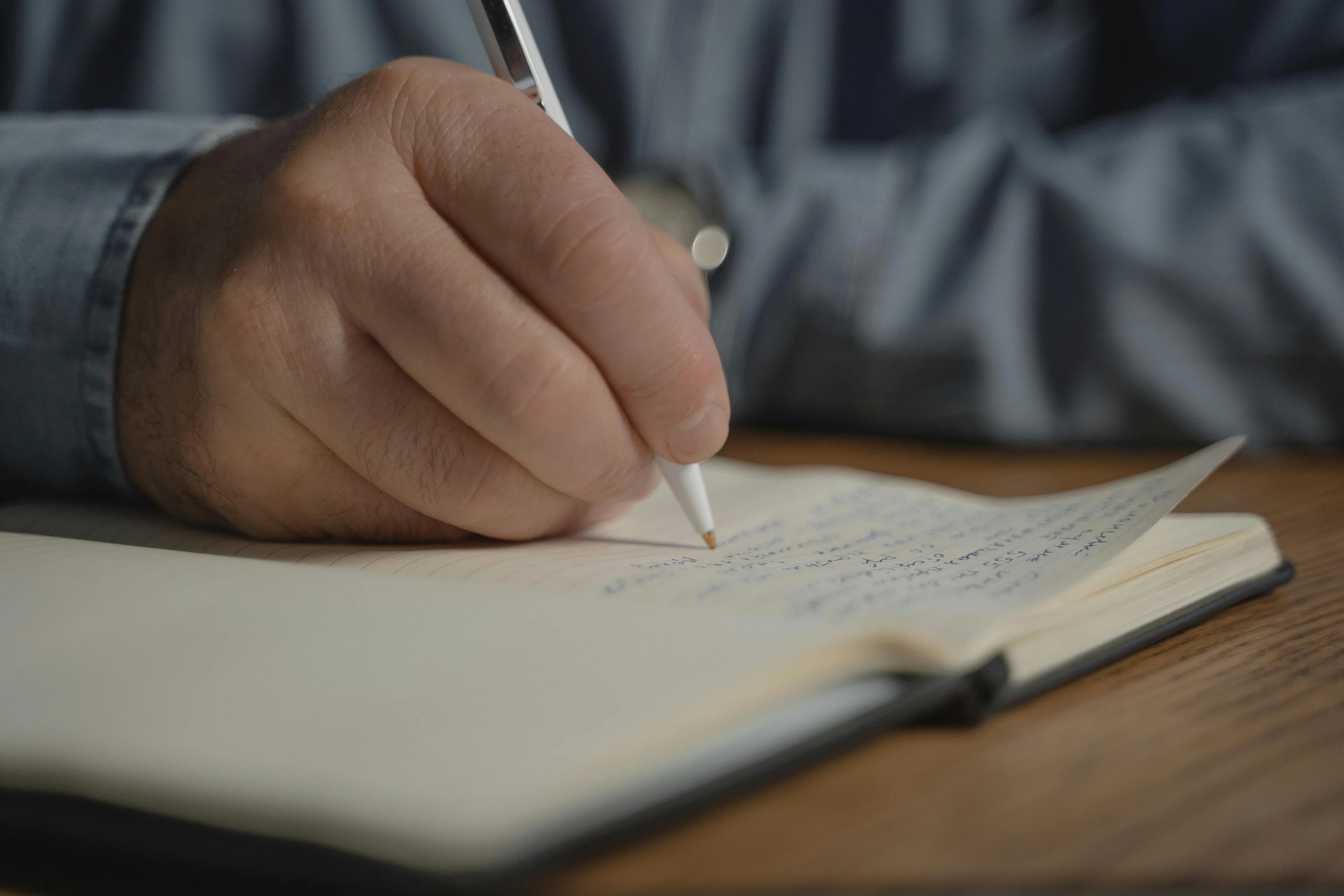 A person's hand in a blue shirt writing in a notebook, engaging in creative writing.