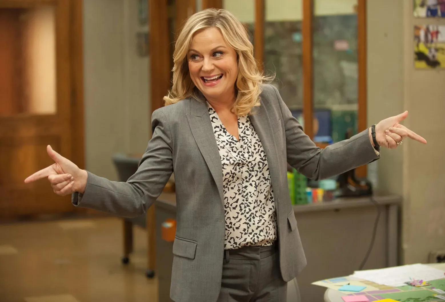 Leslie Knope, a cheerful woman in a gray suit, enthusiastically points to her sides in an office setting. Fictional mentor.