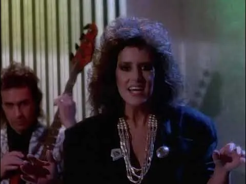 A woman with big 80s hair and a layered necklace sings, with a man playing a bass guitar behind her. Nothing's Gonna Stop Us Now!