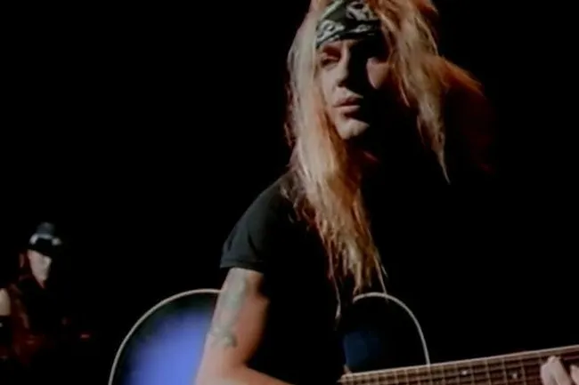Poison's Bret Michaels with long blonde hair and bandana playing acoustic guitar. An '80s power ballad, Every Rose Has Its Thorn.