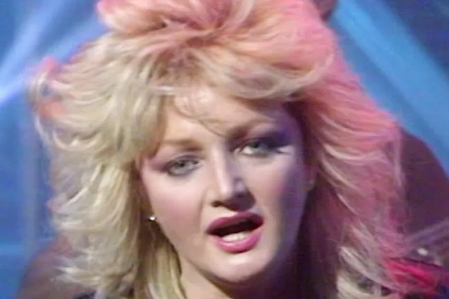 A woman with voluminous blonde and pink hair, heavy eye makeup, and pink lipstick, singing. Evokes an 80s power ballad like Total Eclipse of the Heart.
