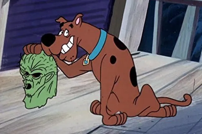 Scooby-Doo cartoon character, a brown dog, holding up a green monster mask with a wide grin.