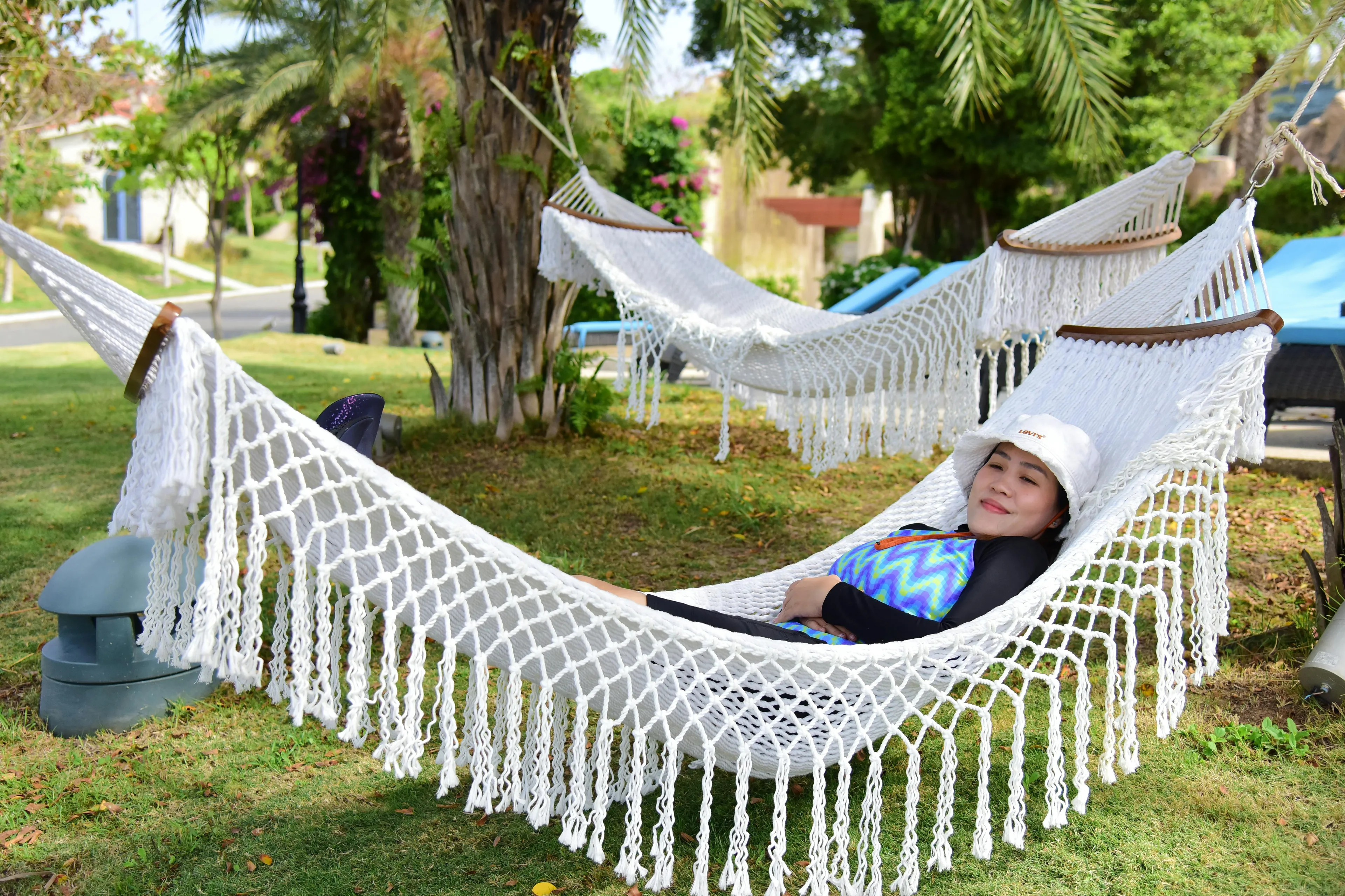 A woman relaxing in a white macrame hammock under palm trees, enjoying a summer day activity, lying in a hammock.