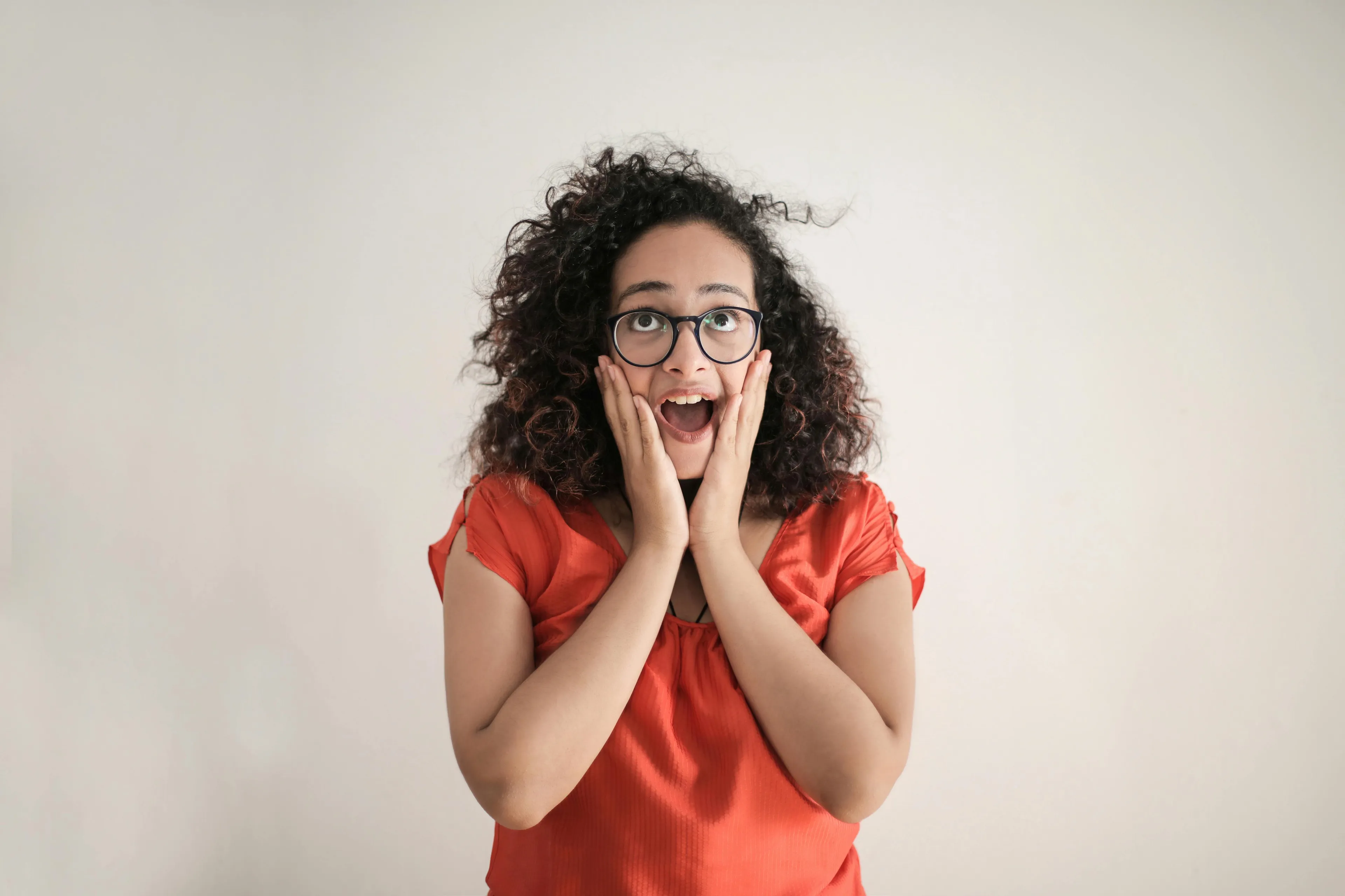 Surprised woman in glasses, hands on cheeks, mouth open, thinking about her self-sabotage pattern.