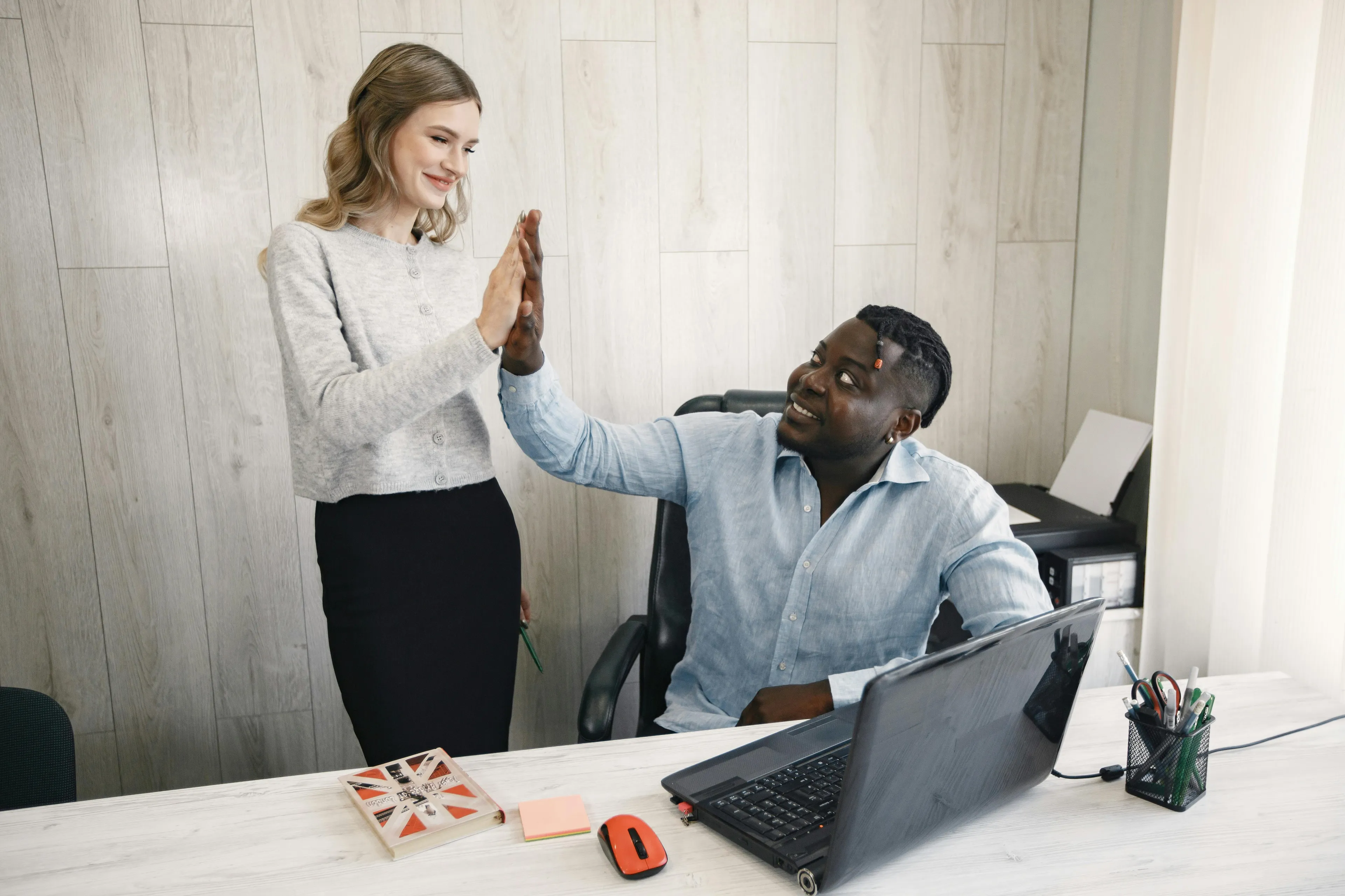 Two colleagues high-fiving at a desk, a woman standing and a man seated. Exploring self-sabotage patterns related to compliments.