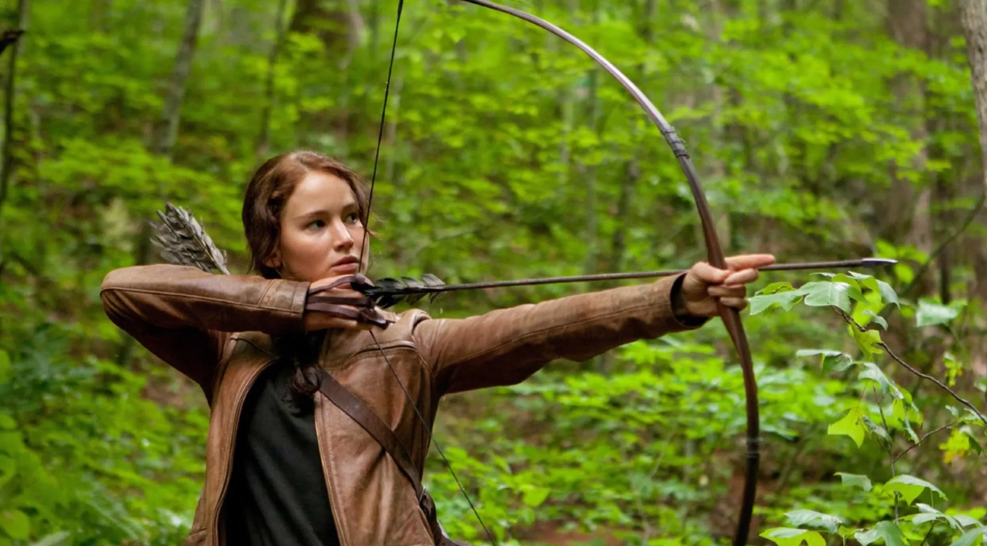 Katniss Everdeen from The Hunger Games, aiming a bow and arrow in a forest. Name the character.
