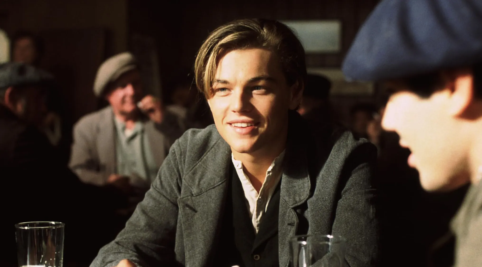 Name The Character: Jack Dawson, smiling in a dimly lit setting with other men. He wears a grey jacket and white shirt.