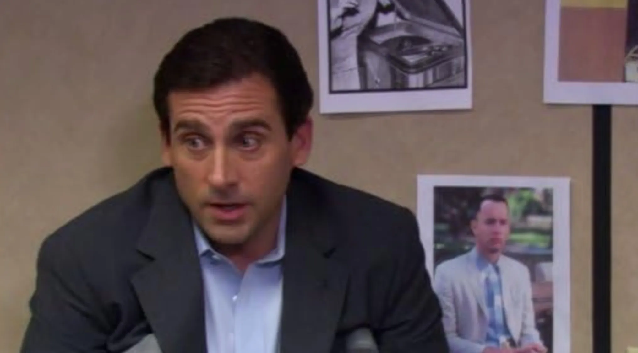 A man, Michael Scott, with wide eyes and open mouth, wearing a dark suit jacket and light blue shirt. Behind him are photos, including one of Forrest Gump. This image helps to Name The Character.