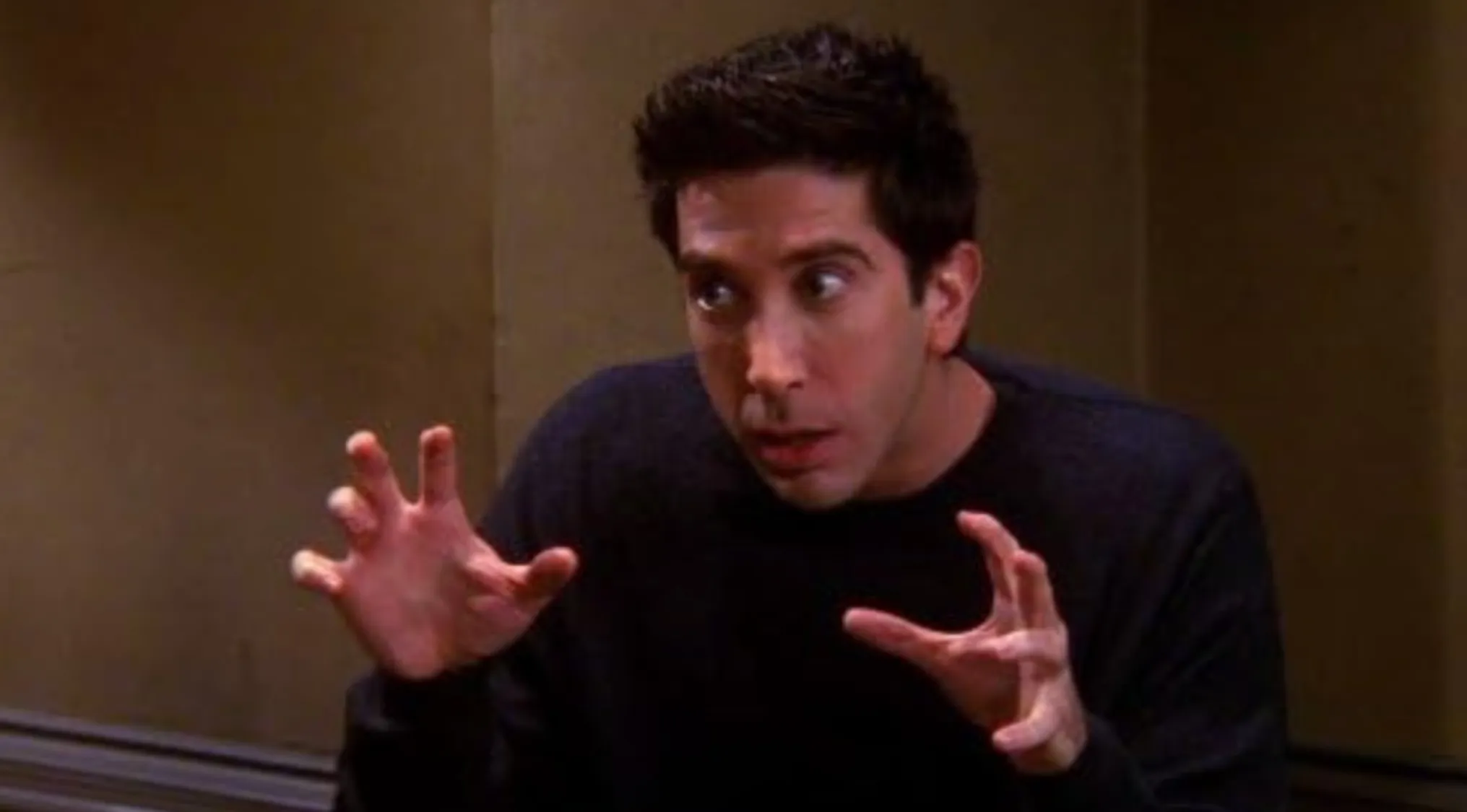 A man with dark hair, a black shirt, and a surprised expression, holding his hands up as if to emphasize something, helps identify the character.