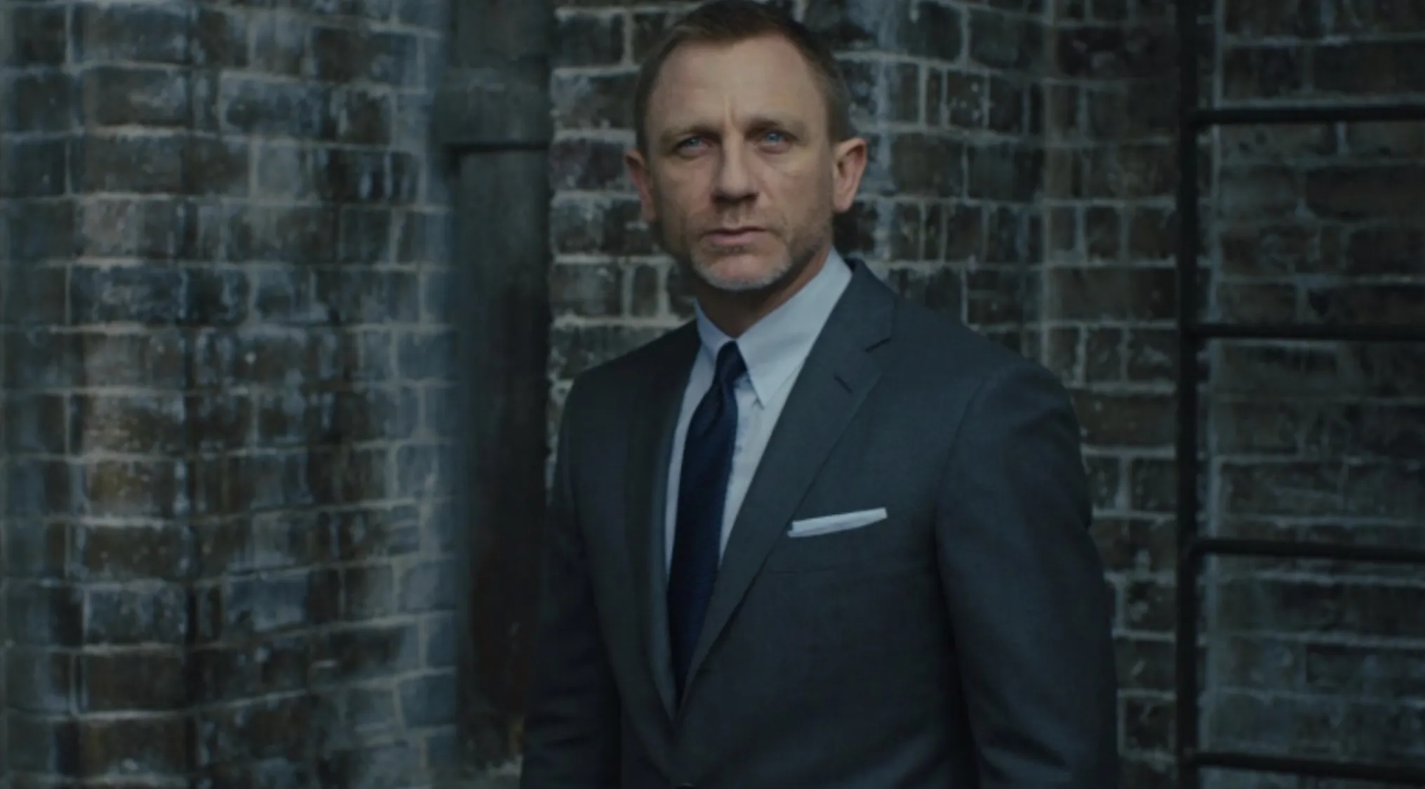 James Bond in a grey suit and blue tie, standing against a dark brick wall, portraying a serious demeanor.