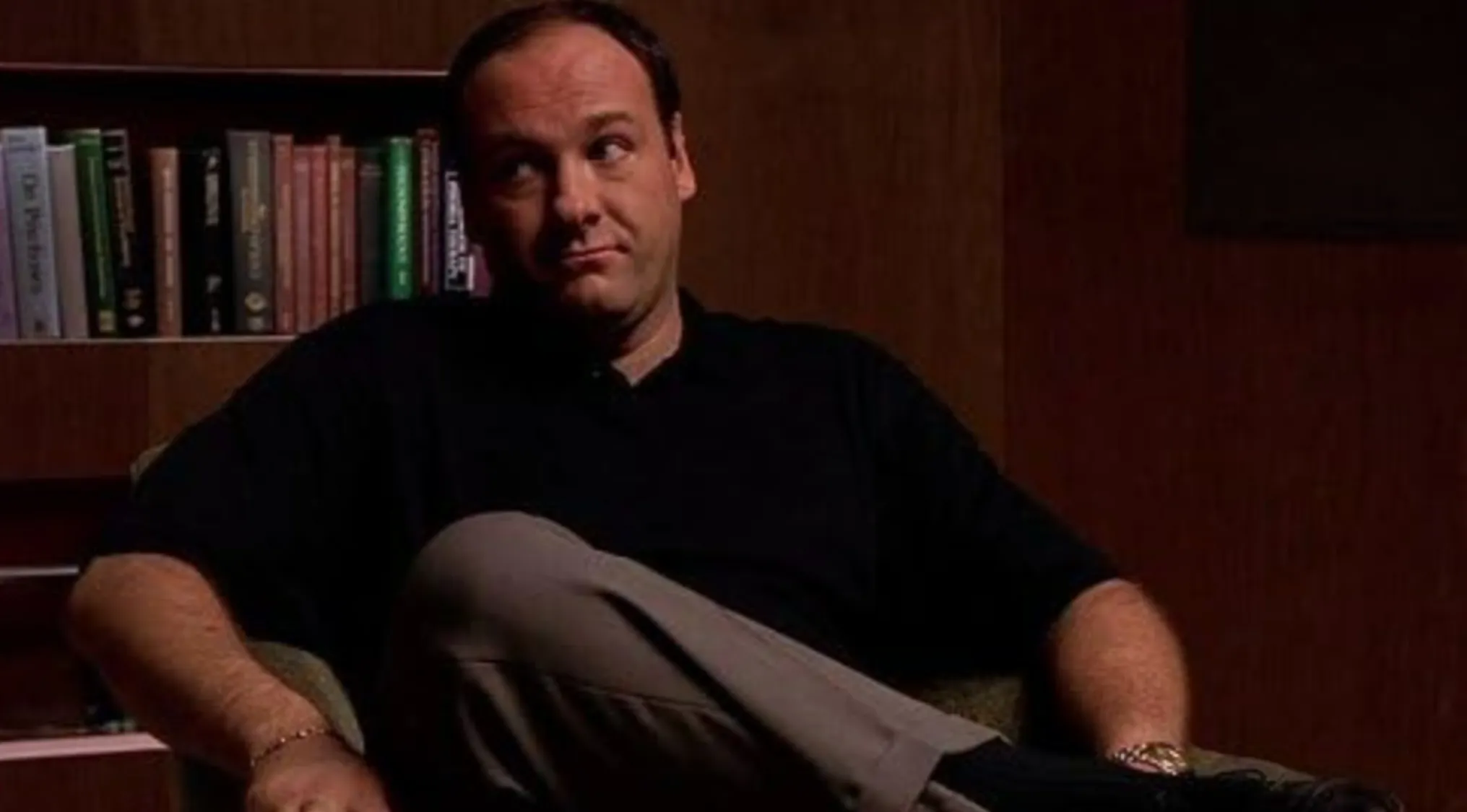 A man in a black shirt and beige pants sits in an armchair, looking sideways. Name the character in this image.