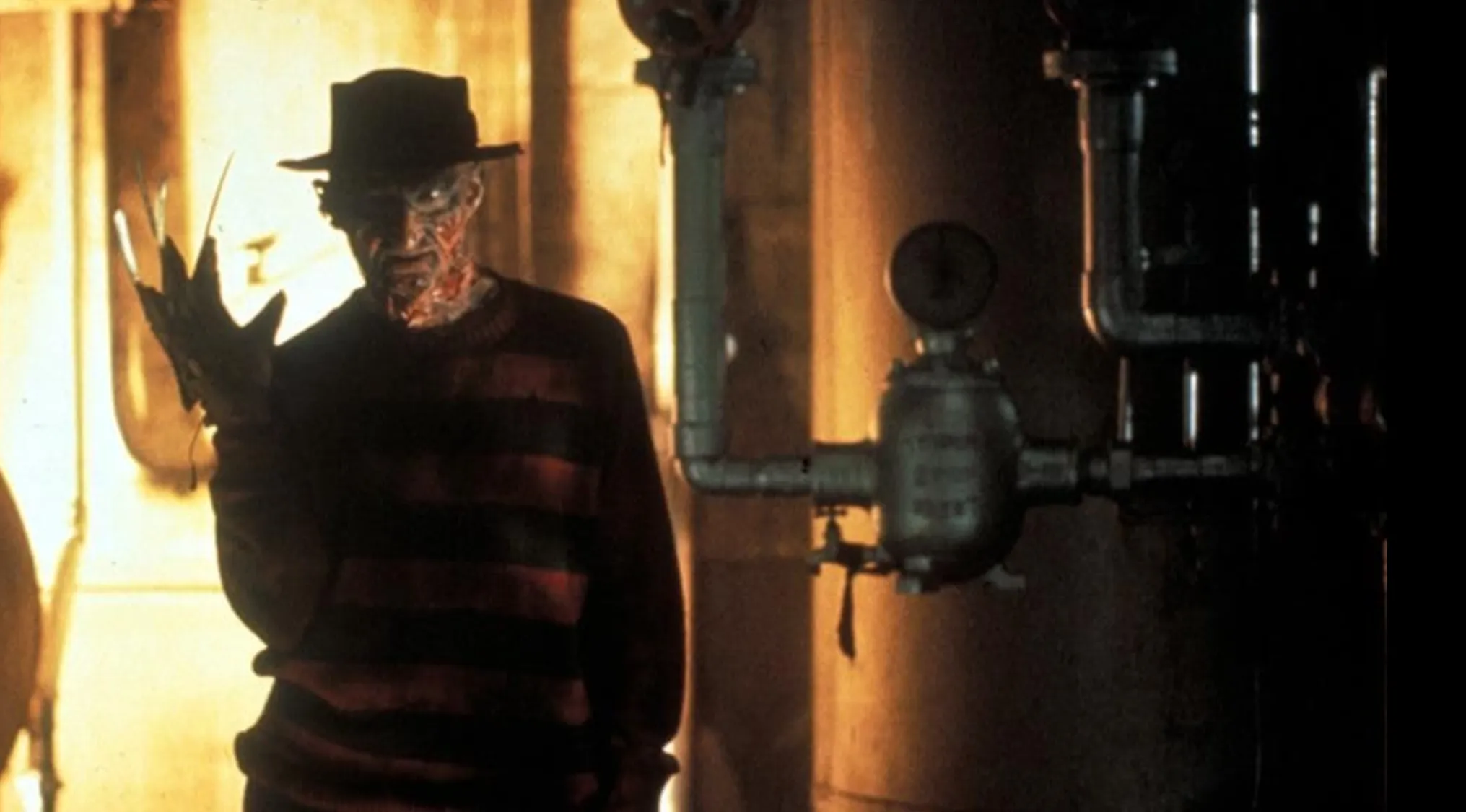 The character Freddy Krueger, wearing his signature hat and striped sweater, with bladed glove raised, standing in a boiler room.
