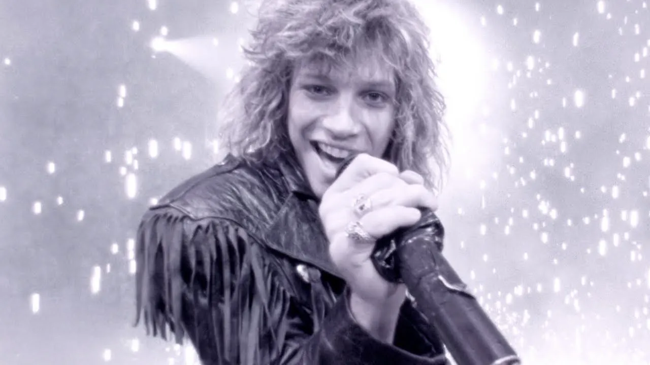 A black and white image of Jon Bon Jovi singing Livin on a Prayer, wearing a fringed leather jacket, mic in hand.