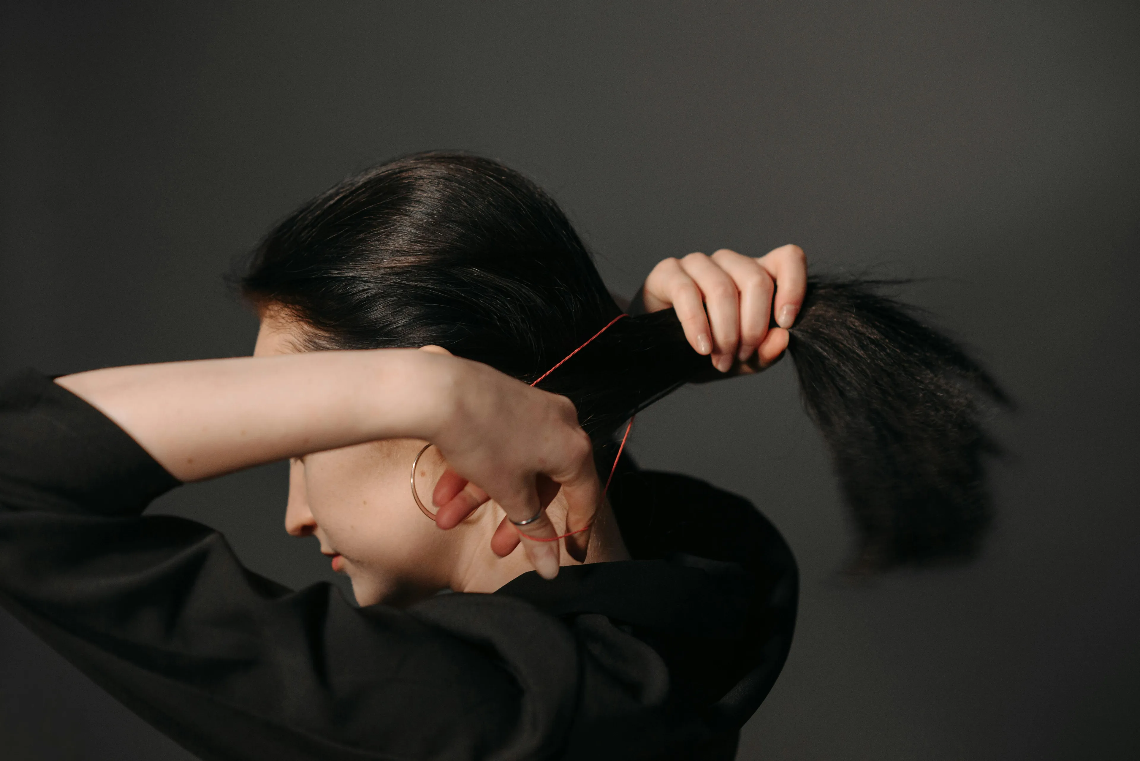 A woman with long dark hair, wearing a black top, pulls her hair back with a red elastic band. Focus on hair protein.