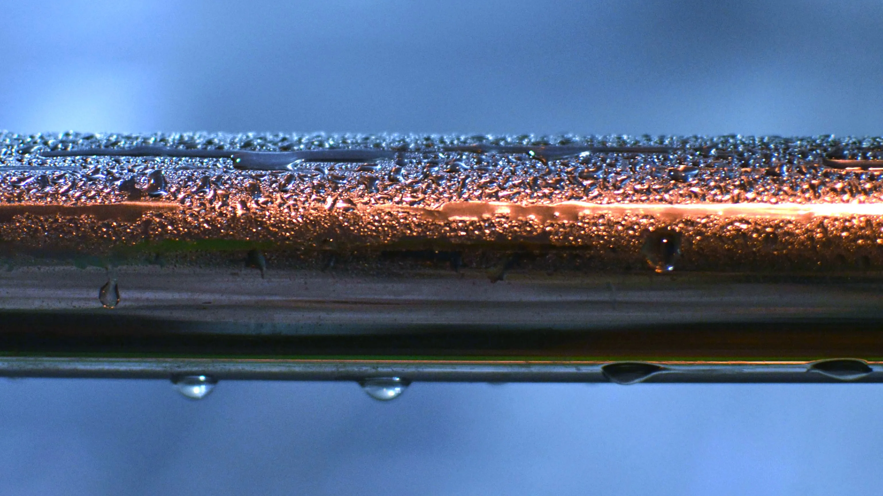 Close-up of a cold metallic pipe with water condensation and droplets, lit by an orange glow. Science type-in trivia.