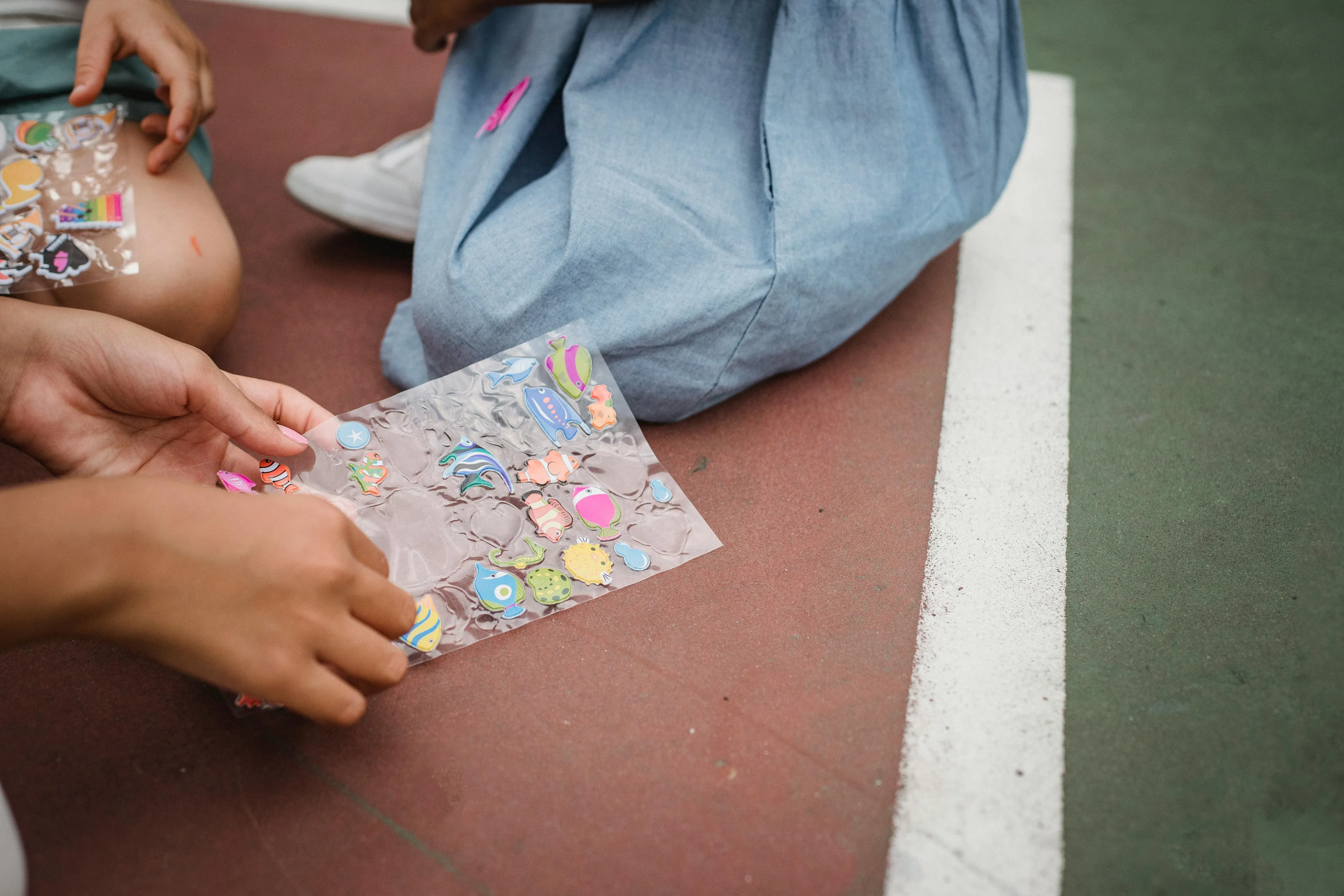 Hands peel colorful fish and sea creature designs from clear sticker sheets. A child in a denim dress sits nearby.