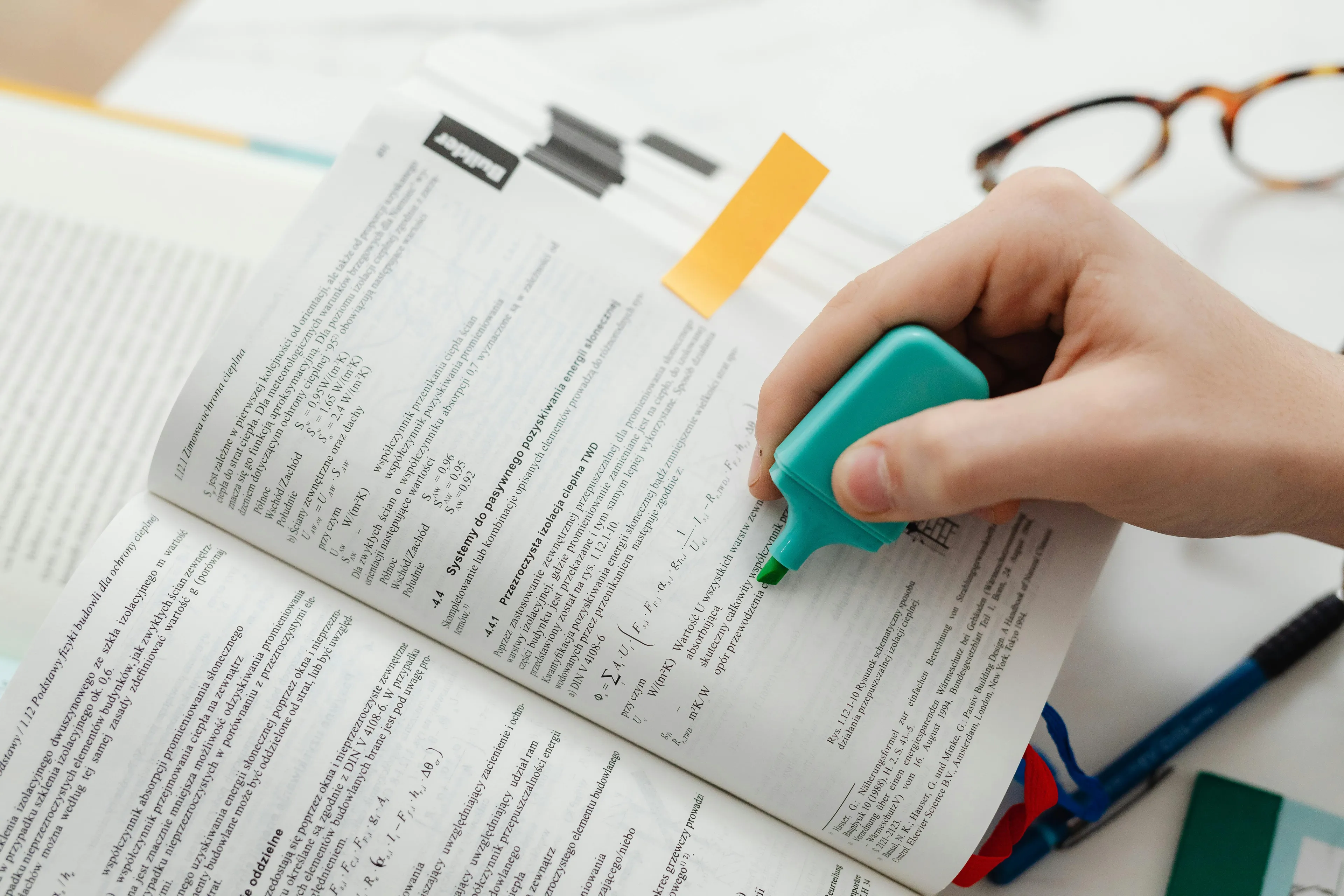 A hand highlighting text in an open book with a turquoise pastel highlighter. Glasses and a pen are nearby.