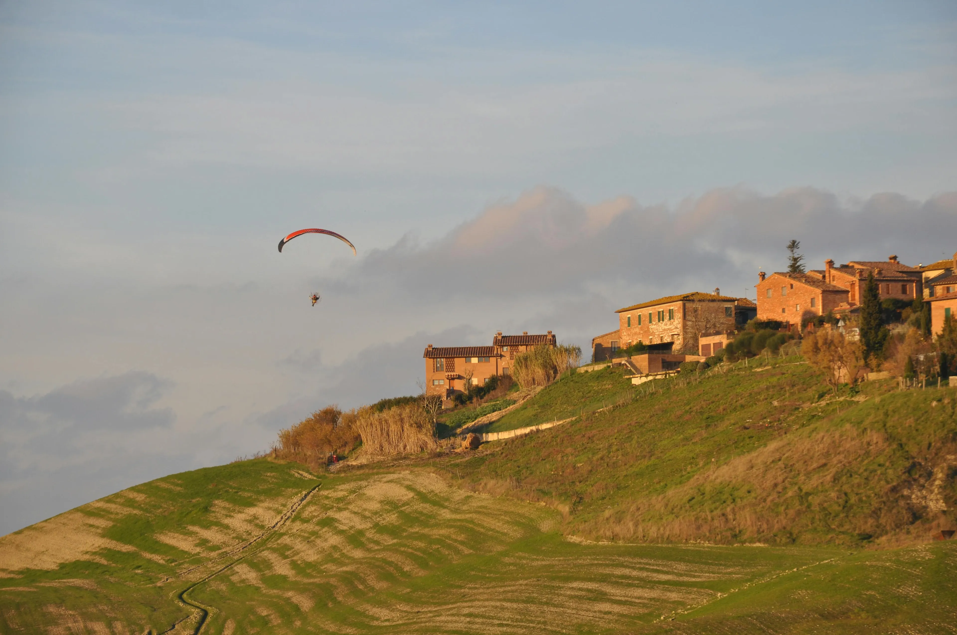 Paraglider soaring over a vibrant green hill with historic buildings in Tuscany, a beautiful travel destination.
