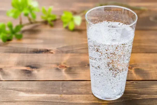 A glass of sparkling water, full of bubbles, sitting on a wooden picnic table with green leaves in the background.