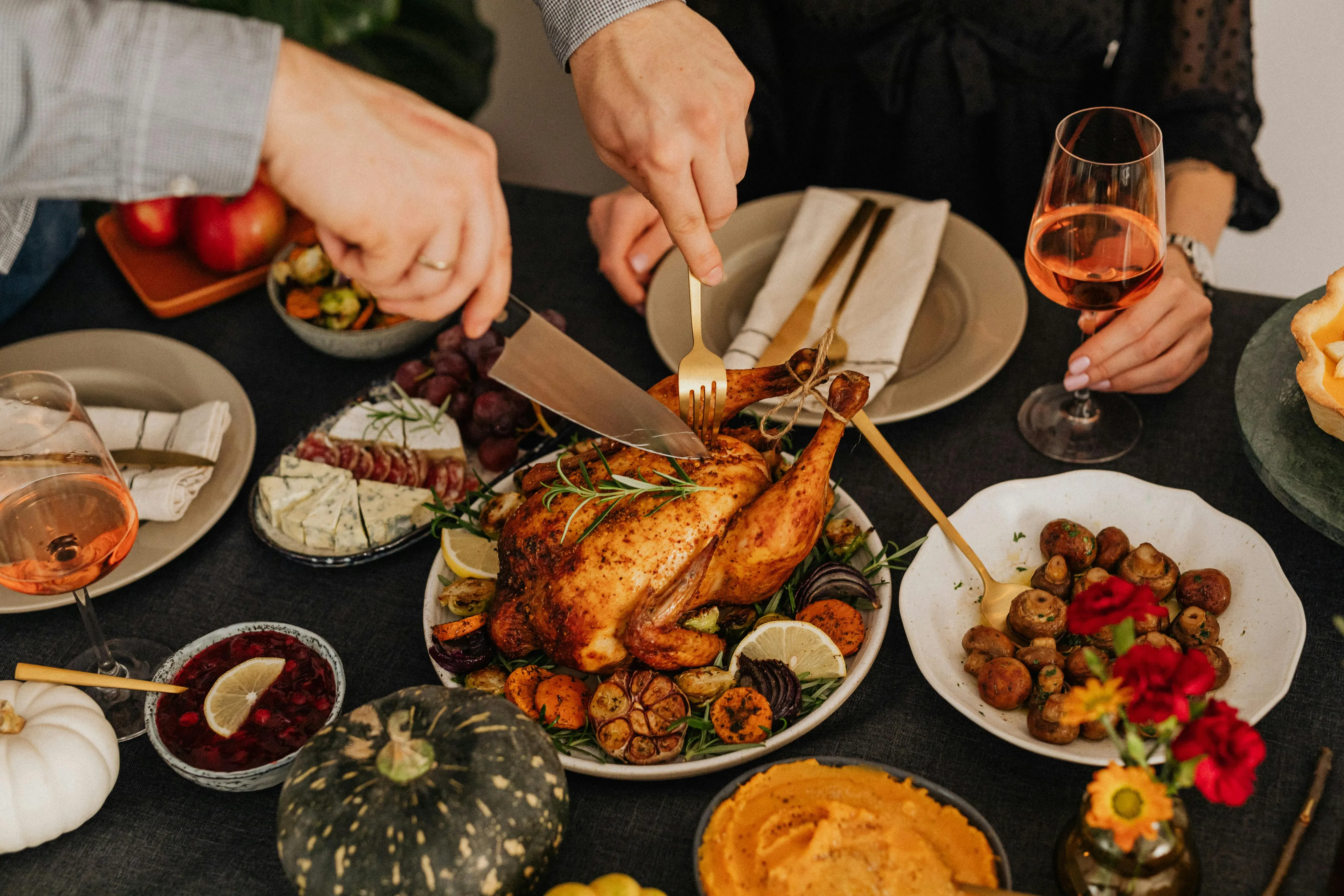 People carving a roasted turkey at a Thanksgiving dinner table, surrounded by festive side dishes and drinks.
