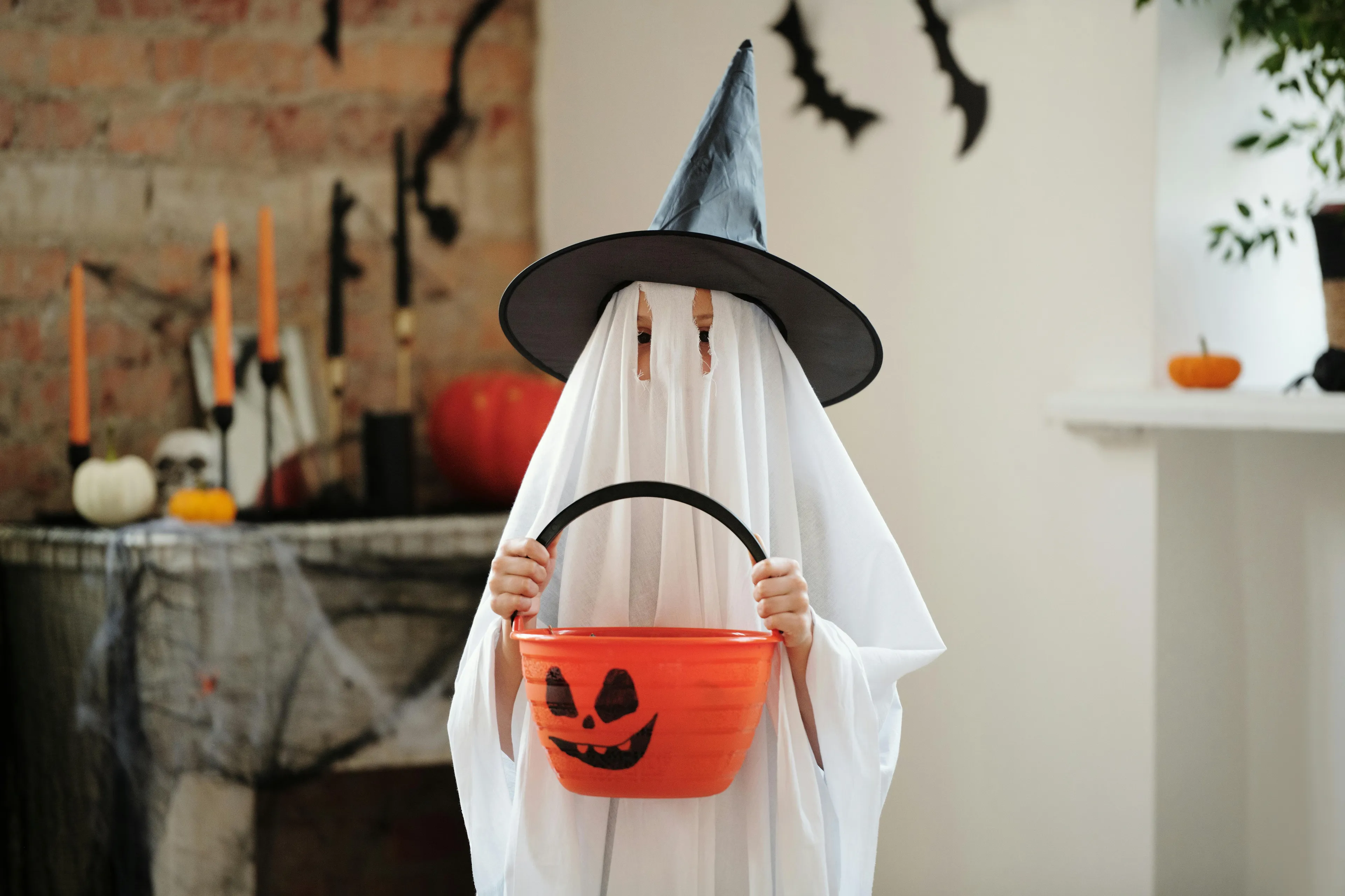 A child dressed as a ghost with a witch hat, holding a pumpkin candy bucket for Halloween. Festive decorations are in the background.